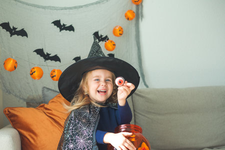 Little cute girl 3 years old in a witch costume with a pumpkin bucket full of sweets celebrates Halloween at home with decorations. Holidays.の写真素材