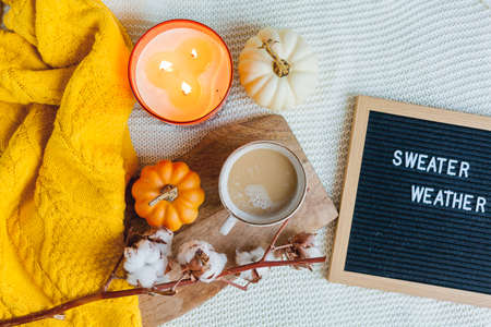 Autumn flatlay with a knitted orange sweater, a cup of hot coffee with milk, a white teapot, a burning candle, a pumpkin, a branch of cotton, letter board.の写真素材