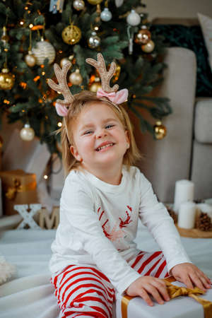 Christmas. Adorable little girl 2 years old with deer horns on her head, in pajamas near the Christmas tree at home. Cozy.の写真素材