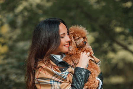 A beautiful young dark-haired woman walks in the park and hugs her poodle dog. Autumn mood. Fall.の写真素材