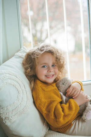 Little cute curly haired girl in knitted pullover sits in front of the window. Cozy home. Warm autumn. Mood.の写真素材