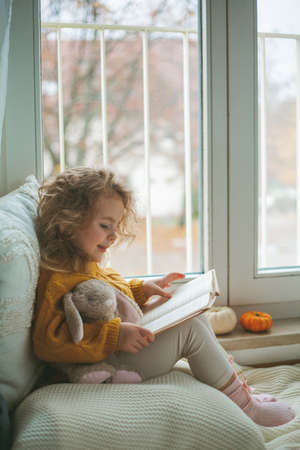 Little cute curly haired girl in knitted pullover sits in front of the window. Cozy home. Warm autumn.の写真素材