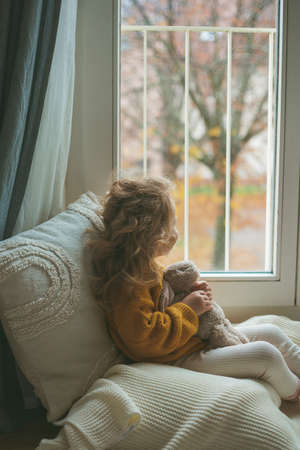 Little cute curly haired girl in knitted pullover sits in front of the window. Cozy home. Warm autumn. Mood.の写真素材