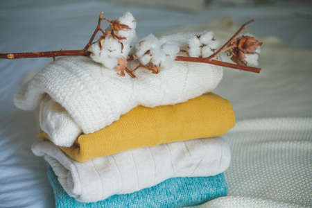 Stack of knitted warm sweaters in wicker rattan basket, plaid, branch of cotton. Autumn mood. Fall.