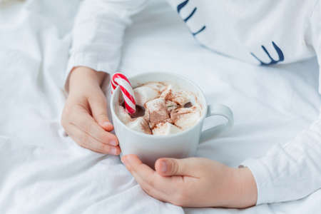 Adorable 6 year old boy in festive pajamas sits by the Christmas tree holding a mug of hot cocoa with marshmallows and lollipop in Christmas decorations. Winter holidays. New Year.の写真素材