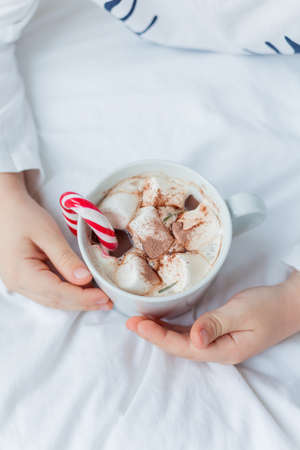 Adorable 6 year old boy in festive pajamas sits by the Christmas tree holding a mug of hot cocoa with marshmallows and lollipop in Christmas decorations. Winter holidays. New Year.の写真素材