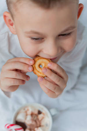 Adorable 6 year old boy in festive pajamas sits by the Christmas tree holding a mug of hot cocoa with marshmallows and lollipop in Christmas decorations. Winter holidays. New Year.の写真素材