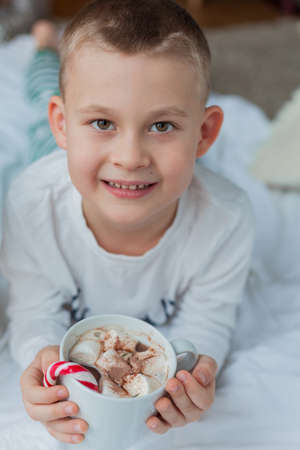 Adorable 6 year old boy in festive pajamas sits by the Christmas tree holding a mug of hot cocoa with marshmallows and lollipop in Christmas decorations. Winter holidays. New Year.の写真素材