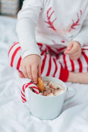 Adorable 2 year old girl in festive pajamas sits by the Christmas tree holding a mug of hot cocoa with marshmallows and lollipop in Christmas decorations. Winter holidays. New Year.の写真素材