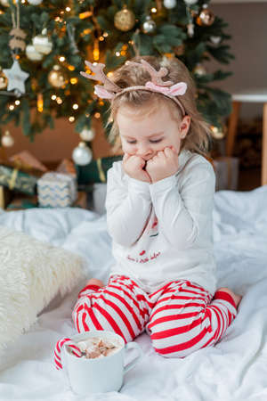 Adorable 2 year old girl in festive pajamas sits by the Christmas tree holding a mug of hot cocoa with marshmallows and lollipop in Christmas decorations. Winter holidays. New Year.の写真素材