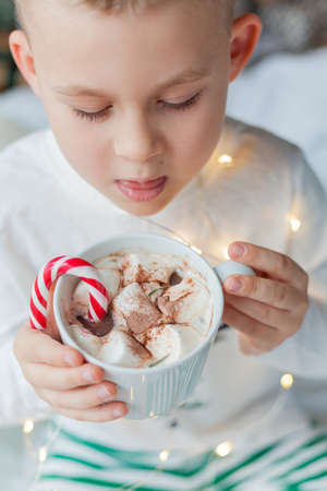 Adorable 6 year old boy in festive pajamas sits by the Christmas tree holding a mug of hot cocoa with marshmallows and lollipop in Christmas decorations. Winter holidays. New Year.の写真素材