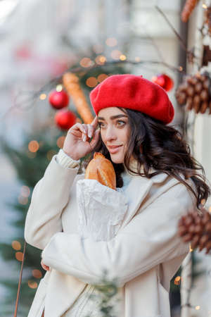 Christmas street style portrait of young beautiful woman in red beret walking in European city for winter holidays. Stylish model with curly hair is doing New Year's shopping. Cozy.の写真素材