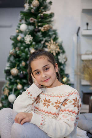 Adorable dark haired little girl 9 years old in pajamas near the Christmas tree. New Year's lights. Winter holidays. Cozy.の写真素材
