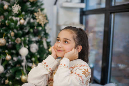 Adorable dark haired little girl 9 years old in pajamas near the Christmas tree. New Year's lights. Winter holidays. Cozy.の写真素材