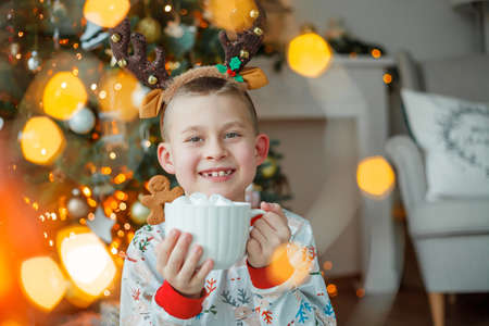 Adorable 7 year old boy in pajamas drinks hot cocoa with marshmallows in white mug with gingerbread man cookies near the Christmas tree. New Year's lights. Winter holidays. Cozy home.の写真素材
