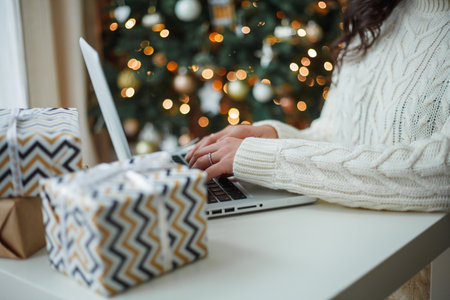 Happy young dark-haired curly woman in a light knitted sweater prepares gifts near the Christmas tree. Stylish model makes online purchases during the winter holidays. New Year. Cozy home.の写真素材