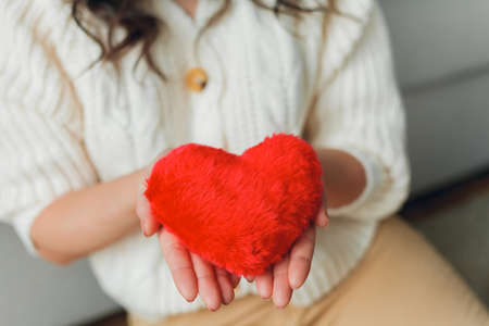 Emotional portrait of 30 year old woman in knitted stylish cardigan. Young beautiful dark haired curly woman holding red plush card heart on valentine's day in her hands. Cozy home.の写真素材