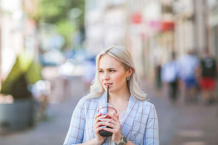 Stylish blonde in casual light clothes with a cocktail in her hands. Portrait of a beautiful fashionable woman on the street of a European city. warm time. shopping. city.の写真素材