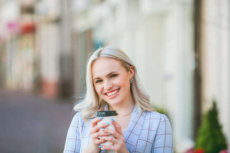 Beautiful young blonde woman drinks coffee from a paper cup and walks around the city. Portrait of a stylish young model in casual clothes outdoors.の写真素材