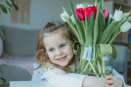 Cute little girl of 3 years old in a cozy knitted sweater hugs a vase with a bouquet of white and pink tulips in a home interior. home comfort.の写真素材