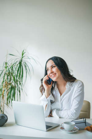 Young businesswoman works at a computer and speaks on the phone. A beautiful dark-haired woman in a white shirt works online in a bright office. Drinking coffee and eating a croissの写真素材