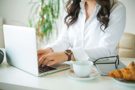 Young businesswoman works at the computer. A beautiful dark-haired woman in a white shirt works online in a bright office. Drinking coffee and eating a croissant. office.の写真素材