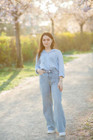 Beautiful young woman of 19 years old in light casual clothes in pastel shades near flowering sakura trees. Spring portrait of model with blue eyes. Spring.の写真素材