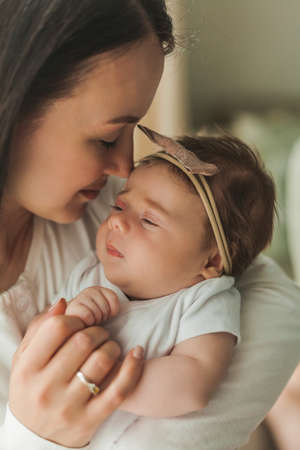 Young beautiful dark-haired mother holds her newborn daughter in her arms in a cozy home. family portrait. motherhood. Young woman kisses her 2 month old baby. home.の写真素材