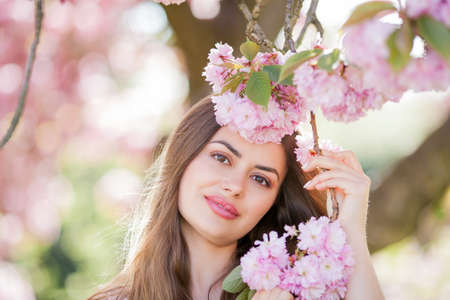 Spring portrait of a young beautiful woman in pink blossoms. Young beautiful model with long hair, in a wicker hat and a light dress near the cherry blossoms. Spring.の写真素材