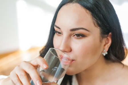 A young beautiful dark-haired woman in a knitted cardigan drinks clean water from a glass. Cozy house model portrait. health. water.の写真素材