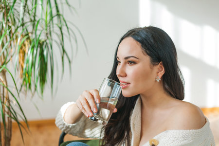 A young beautiful dark-haired woman in a knitted cardigan drinks clean water from a glass. Cozy house model portrait. health. water.の写真素材