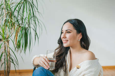A young beautiful dark-haired woman in a knitted cardigan drinks clean water from a glass. Cozy house model portrait. health. water.の写真素材