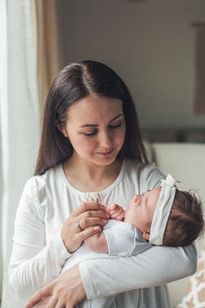Close portrait of a little cute dark-haired newborn girl in a white suit in the arms of a young mother. motherhood. parent. Healthy sleep. home.の写真素材