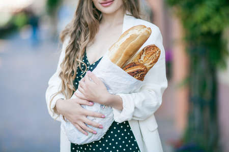 Young beautiful long-haired woman 25 years old in a black and white dress buys a French baguette in the center of a European city. fresh bakery. Stylish model walking around the city. Summer.の写真素材