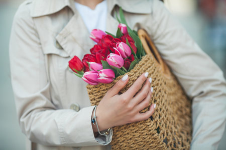 Spring portrait of a young beautiful happy woman 28 years old with long well-groomed hair holds a wicker bag in her hands with a bouquet of tulips on a city street. Stylish model iの写真素材