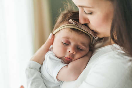 Close portrait of a little cute dark-haired newborn girl in a white suit in the arms of a young mother. motherhood. parent. Healthy sleep. home.の写真素材