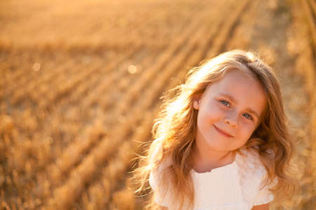 Adorable little curly girl 4 years old in a white dress in the sun at sunset in a mowed field of wheat. happy child outside. Walk. warm summer.の写真素材