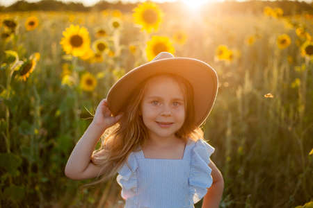 Adorable little girl 4 years old with curly long blond hair in a blue sundress and hat in a sunflower field. Summer portrait of a happy child.の写真素材