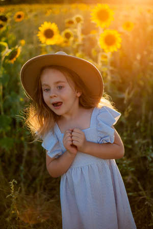 Adorable little girl 4 years old with curly long blond hair in a blue sundress and hat in a sunflower field. Summer portrait of a happy child.の写真素材