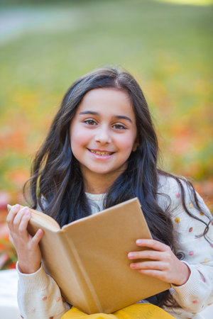 A pretty dark-haired 10-year-old girl in a knitted sweater reads a book sitting in a clearing in an autumn park. Picnic in nature. Happy child is learning. fall.の写真素材