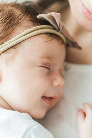 Close portrait of a little cute dark-haired newborn girl in a white suit in the arms of a young...