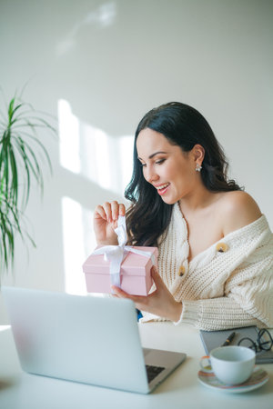 Young beautiful dark-haired woman of 34 years old in knitted sweater is talking on video link and holding pink gift box in her hands. Valentine's Day. Model received gift. holidays.の写真素材