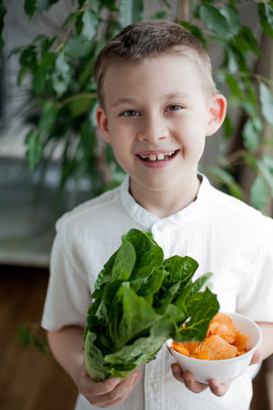 Health food. Cute little boy of 7 years old holds in one hand green lettuce leaves in the other hand plate with greasy chips. Eco.の写真素材