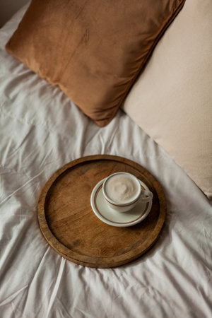 Hot mug of cappuccino on wooden tray on the bed, breakfast. Cozy house. Beige natural colors.の写真素材