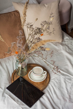Cozy breakfast in bed. Cup of hot cappuccino on wooden tray, bouquet of dried flowers in glass vase,...