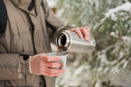 Portrait of young man in warm clothes in winter forest. Drinking hot tea outdoors from flask....
