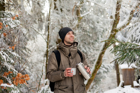 Portrait of young man in warm clothes in winter forest. Drinking hot tea outdoors from flask. Hiking.の写真素材