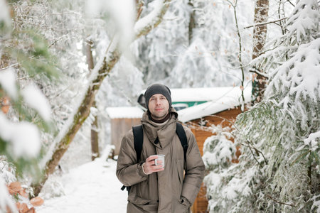 Portrait of young man in warm clothes in winter forest. Drinking hot tea outdoors from flask. Hiking.の写真素材