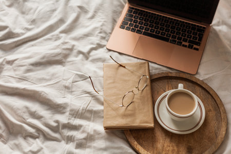 Cup of hot cappuccino, a book, glasses, a laptop on the bed. Work online at home. home office.の写真素材