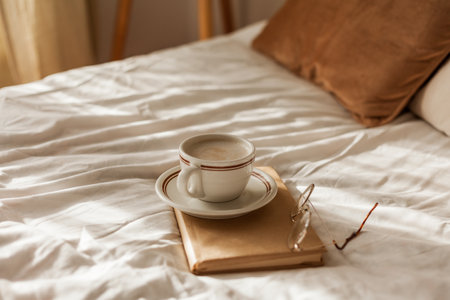 A cup of hot cappuccino, a book, glasses on the bed. Breakfast. Cozy house. decor.